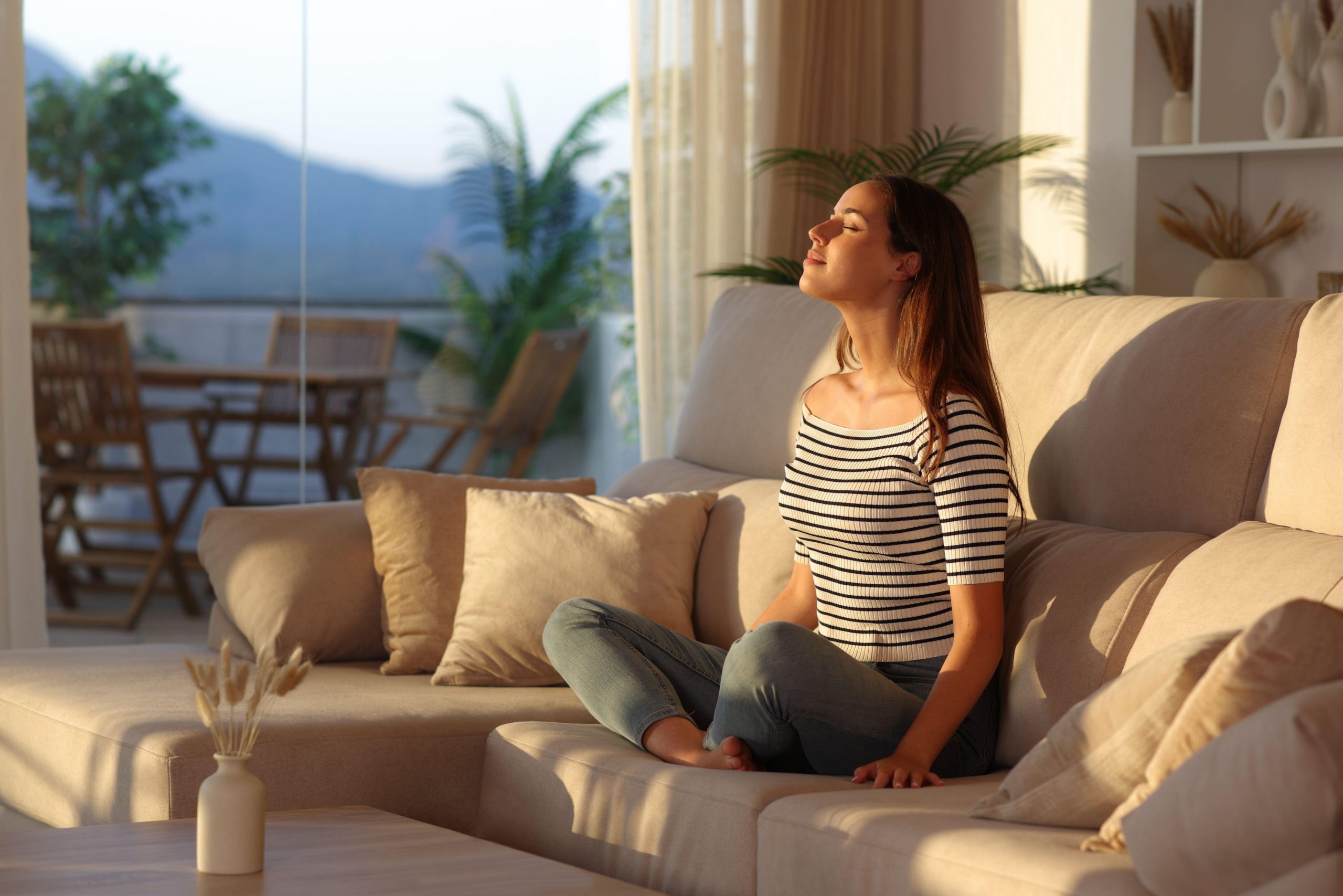 Ženska sedi na kavču po turško, izvaja dihalno vajo, je sproščena in umirjena