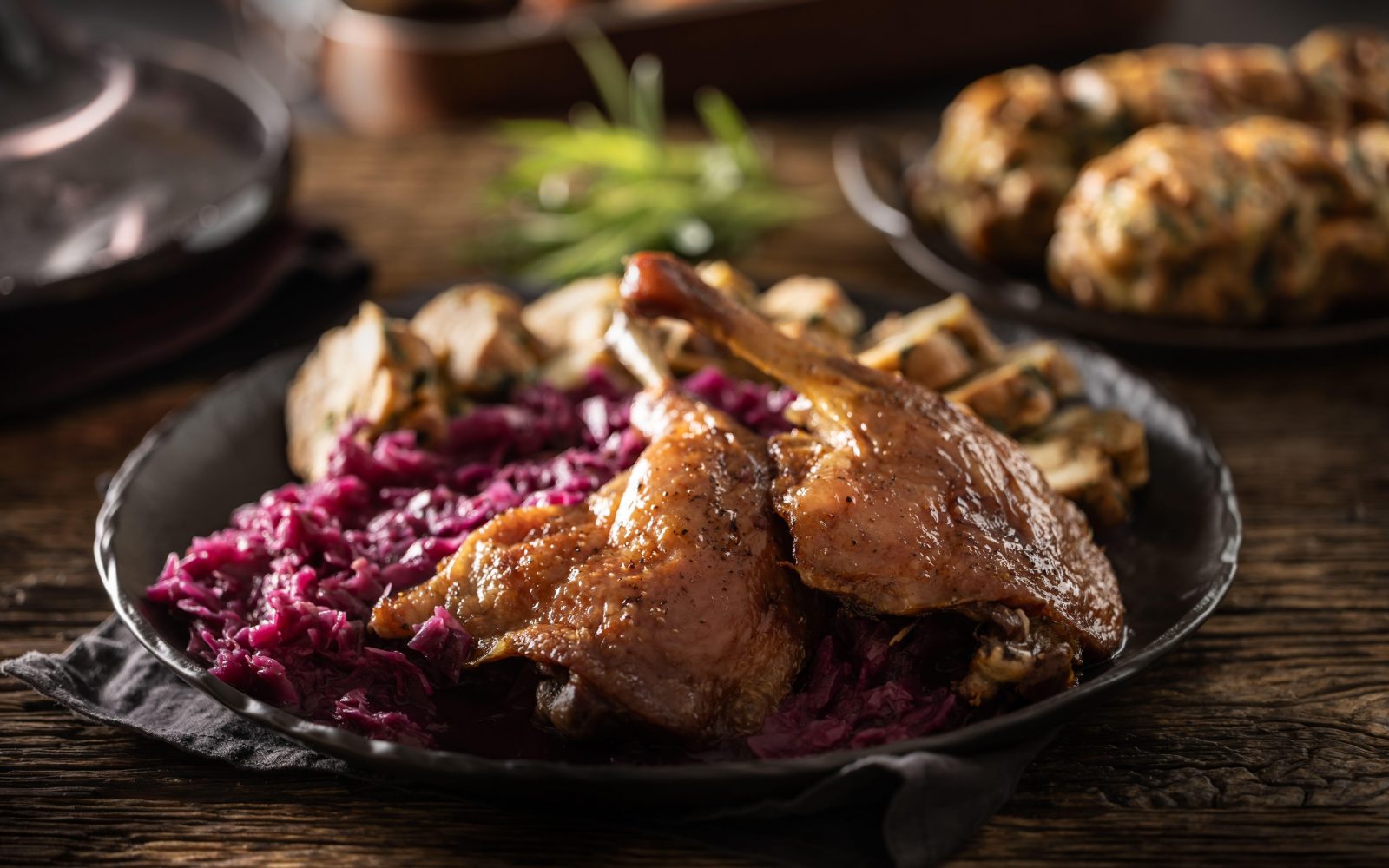 Slika mize ob martinovi pojedini - pečena goska, dušeno rdeče zelje, mlinci in kruhovi cmoki na temnem krožniku, v ozadju vino in druge jesenske priloge