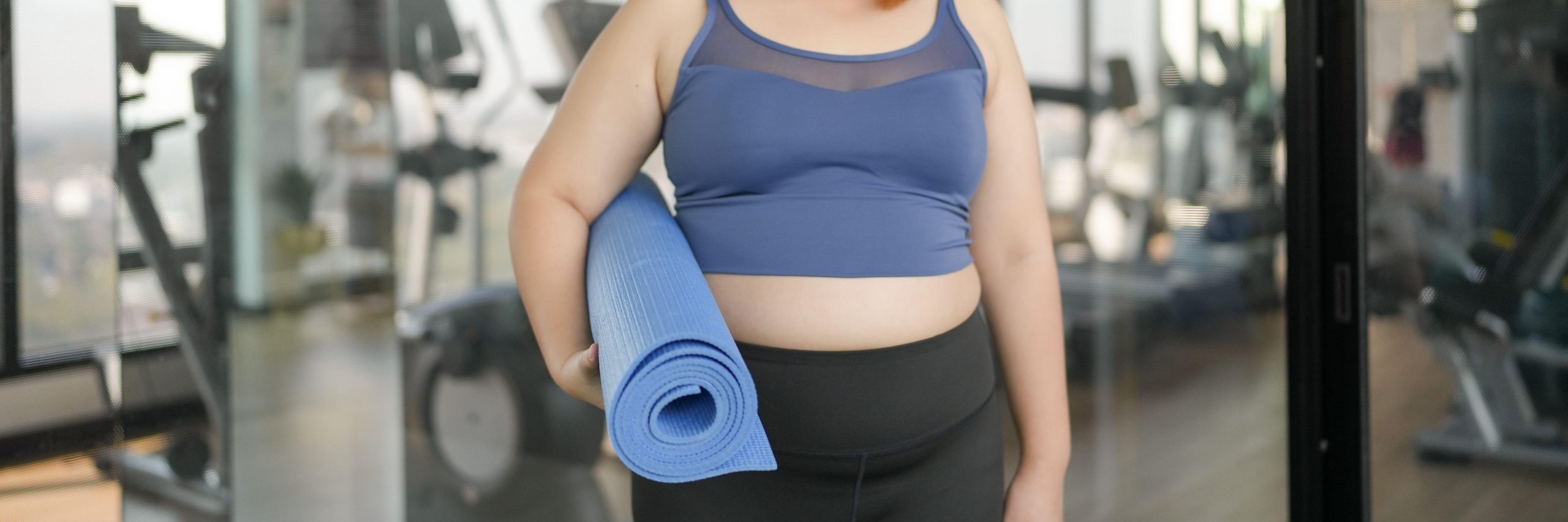 Debelejša ženska v fitnesu v roki drži podlogo za jogo - vadba za znižanje telesne teže