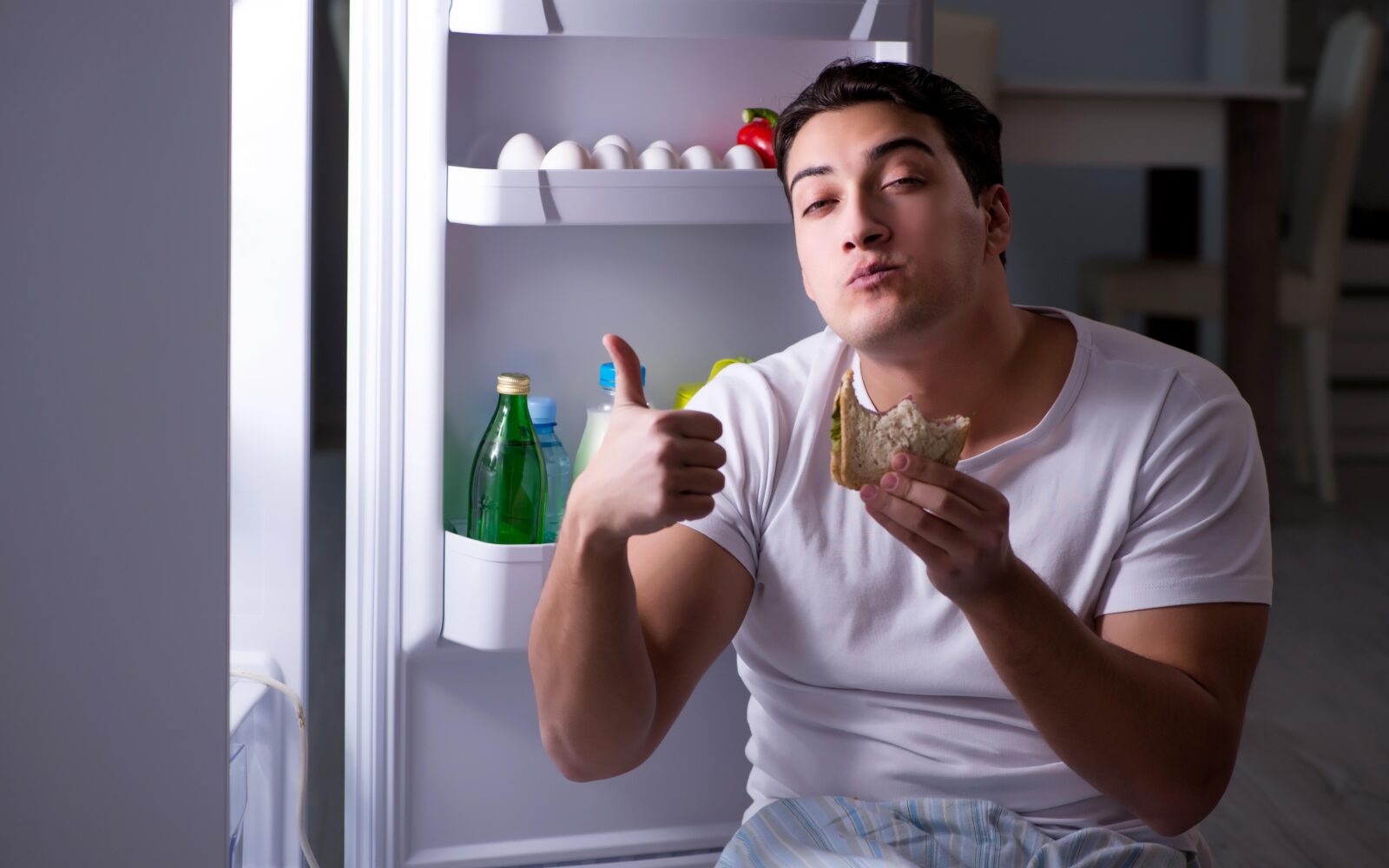 Moški v nočnem času sedi pred hladilnikom in je hrano