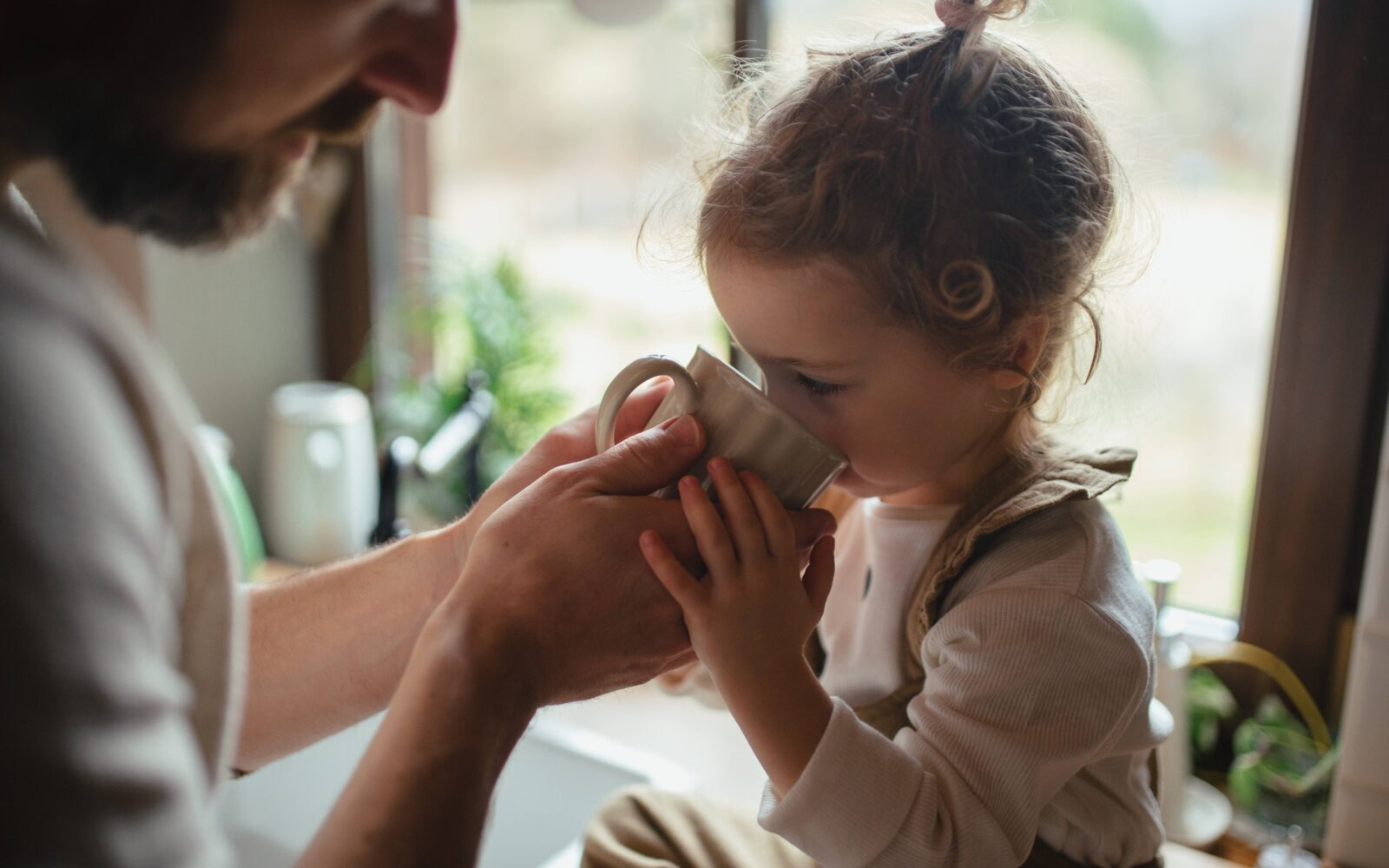 Prehlajena deklica z bolečim grlom sedi na kuhinjskem pultu, očka pa ji iz skodelice pomaga piti topel čaj za blaženje simptomov.