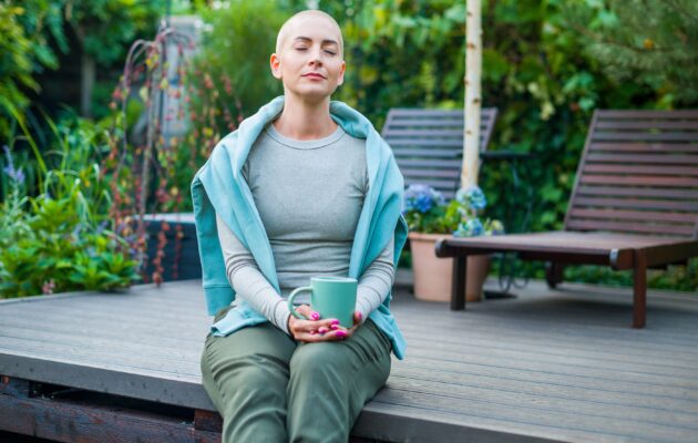 Ženska z rakavo boleznijo je izgubila lase. Sedi na podestu pred hišo, v rokah drži skodelico.