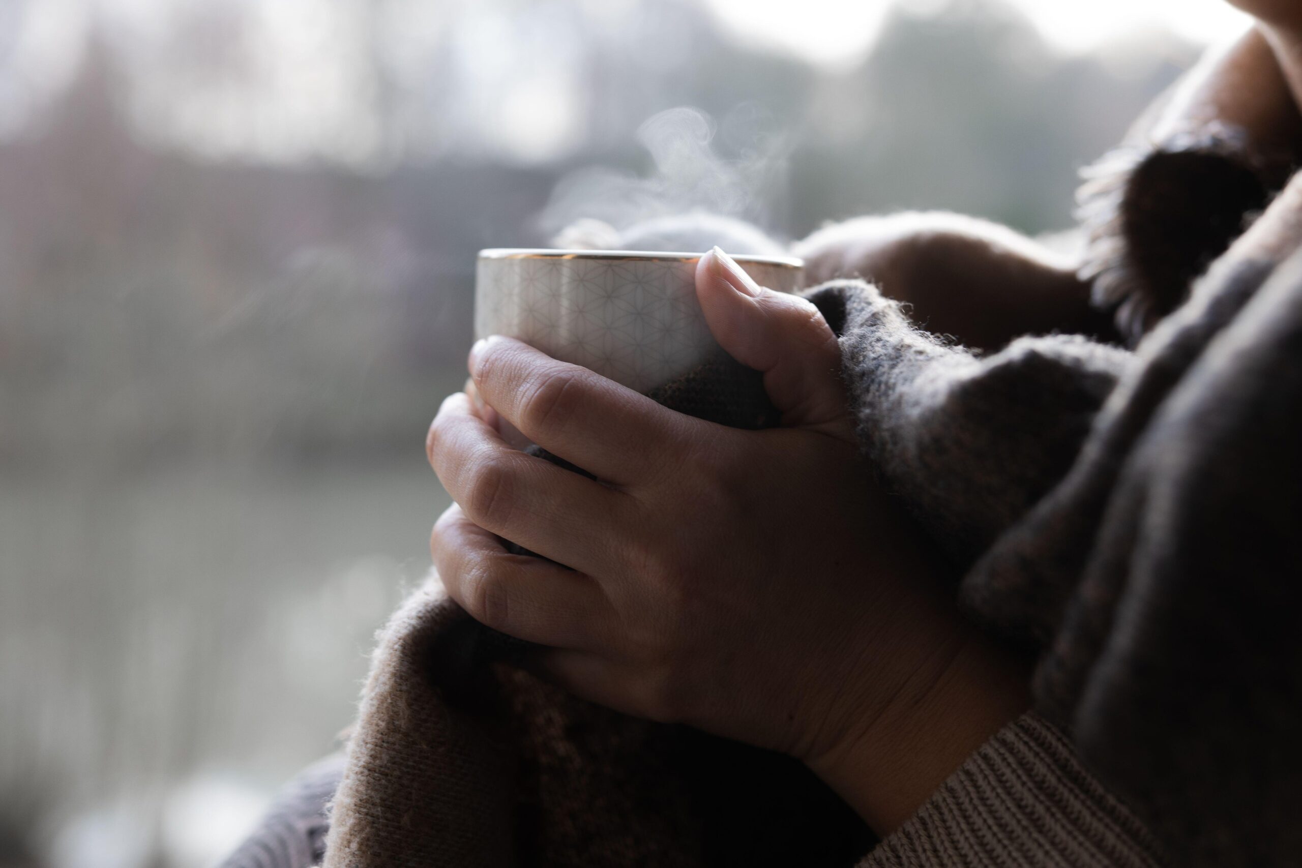 Toplo oblečena ženska sedi pri oknu, pije čaj in skozi okno gleda zimsko idilo.