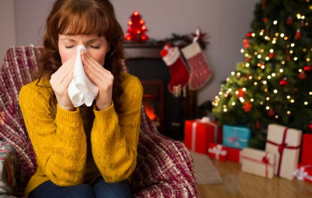 Ženska v rumenem puloverju sedi na kavču, in si briše izcedek iz nosu. Za njo je okrašena novoletna smrečica, pod katero so darila
