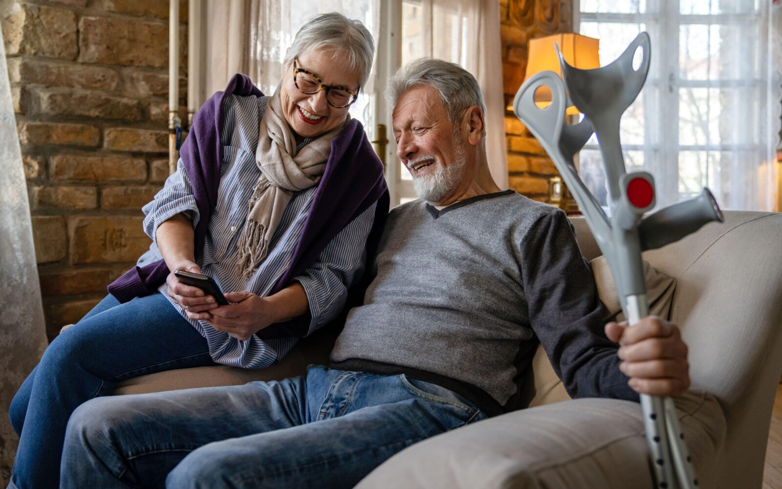 Starejša ženska in starejši moški z berglami sedita na kavču, gledata v telefon in se smejita
