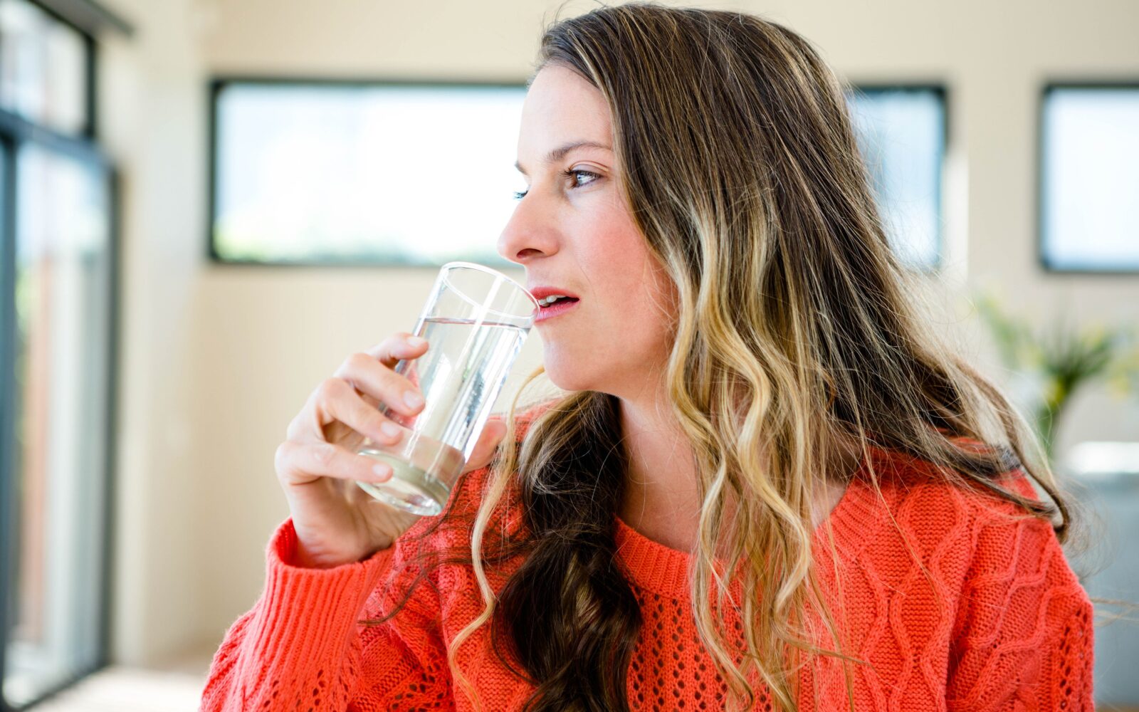 Ženska z dolgimi lasmi, oblečena v oranžni pulover pije vodo