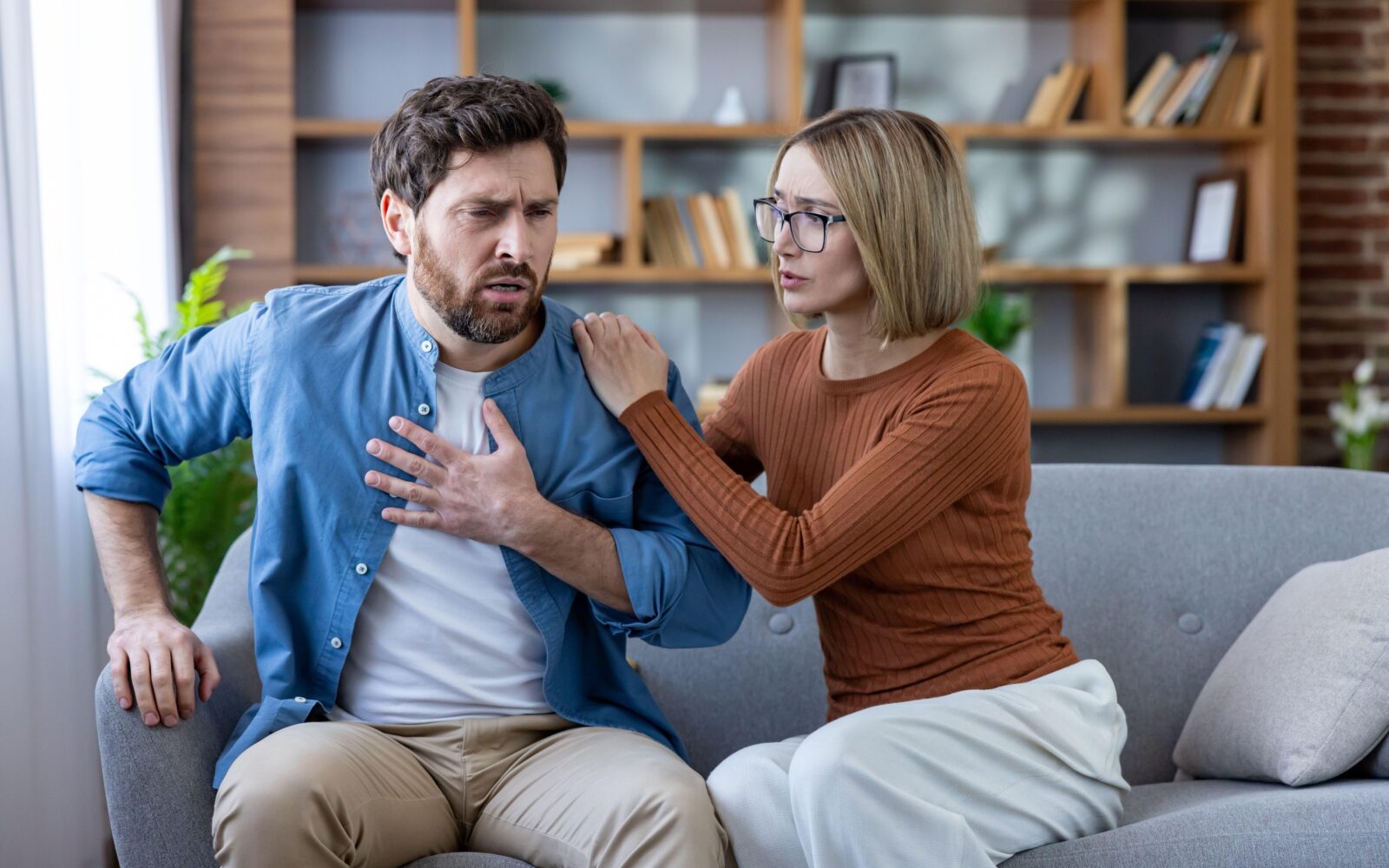 Moški in ženska sedita na kavču, moški j zadihan, občuti tiščanje v prsih, ženska ga zaskrbljeno gleda