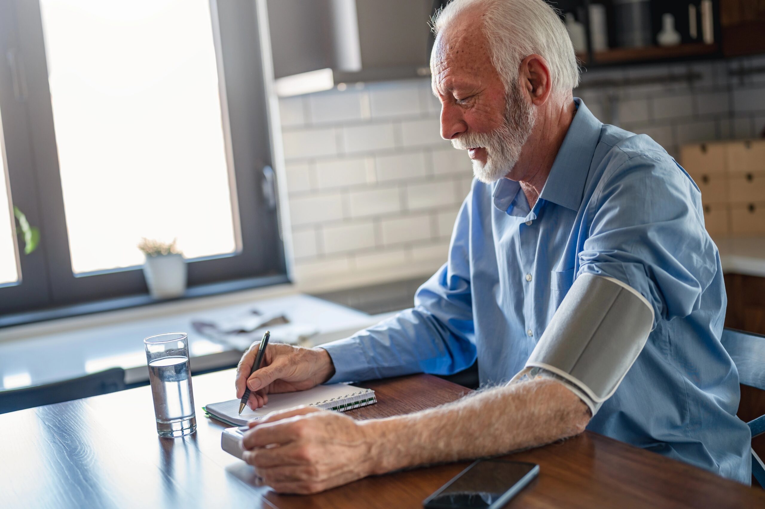 Starejšo moški si doma meri krvni tlak na levi roki, z desno pa piše izmerjene meritve