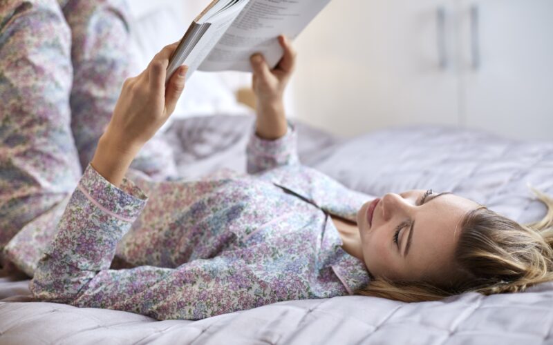 Mlada ženska leži na postelji, oblečena v pižamo in bere knjigo - preprosta aktivnost s številnimi pozitivnimi učinki za telo in možgane