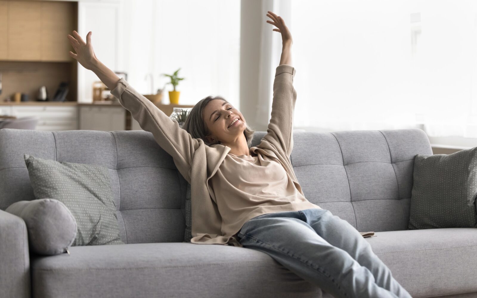 Sproščena ženska sedi na kavču, roke ima dvignjene v zrak, je vesela in nasmejana