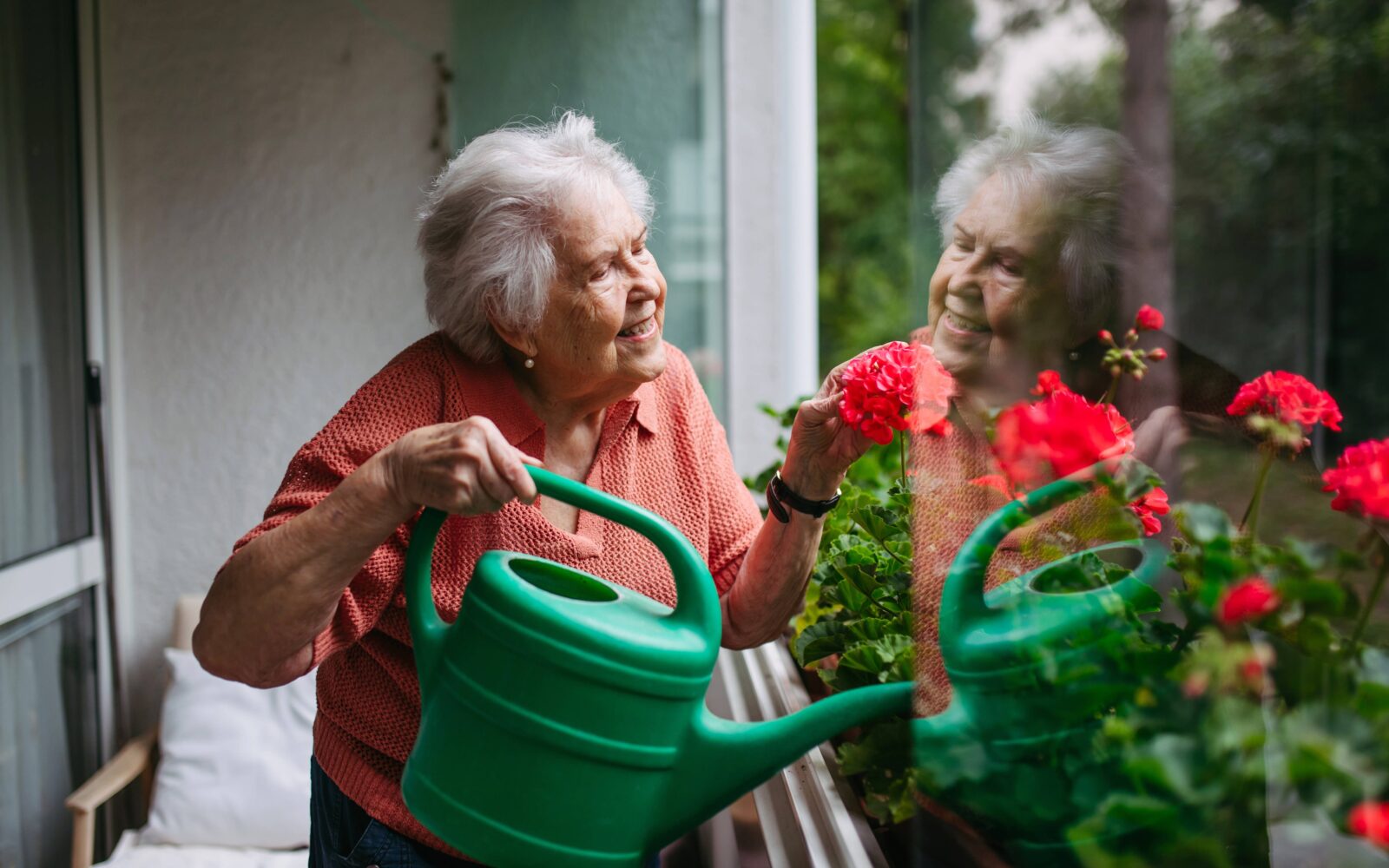 Starejša ženska z demenco zaliva rože na balkonu in ohranja samostojnost, dostojanstvo ter občutek vrednosti pri vsakdanjih opravilih.