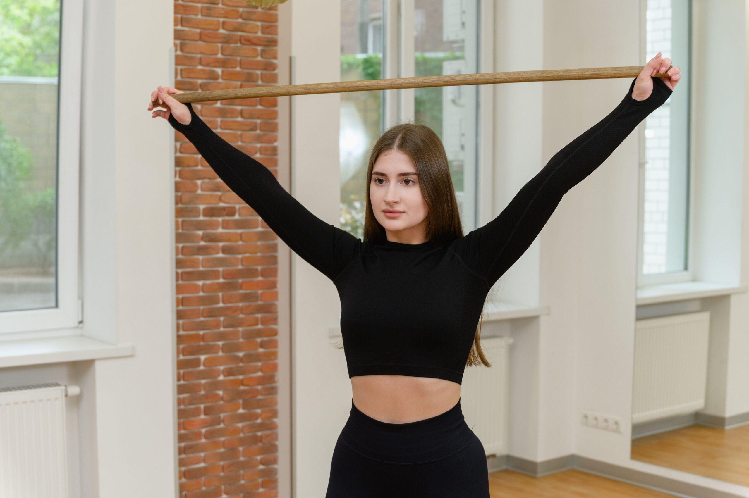 Mlada ženska v črnih športnih oblačilih izvaja vajo za razteg ramen s pomočjo palice.