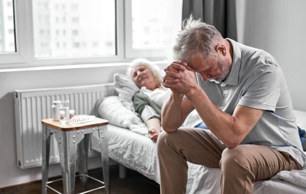Svojec v stiski ob postelji osebe z demenco v bolnišnici, simbol nenadne spremembe stanja in delirija