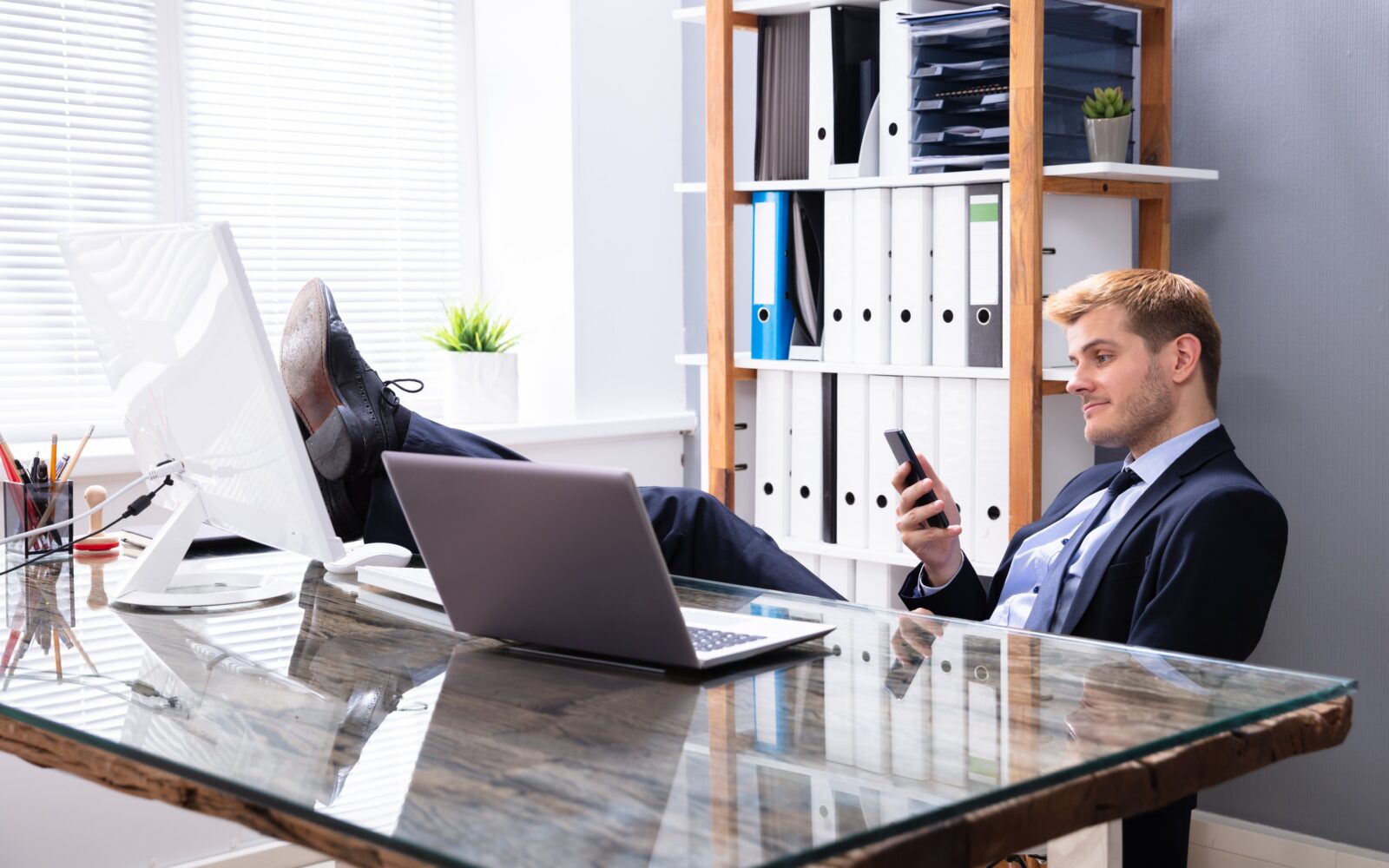 Moški v poslovni obleki sedi za mizo in namesto da bi delal, ima noge na mizi in gleda v telefon