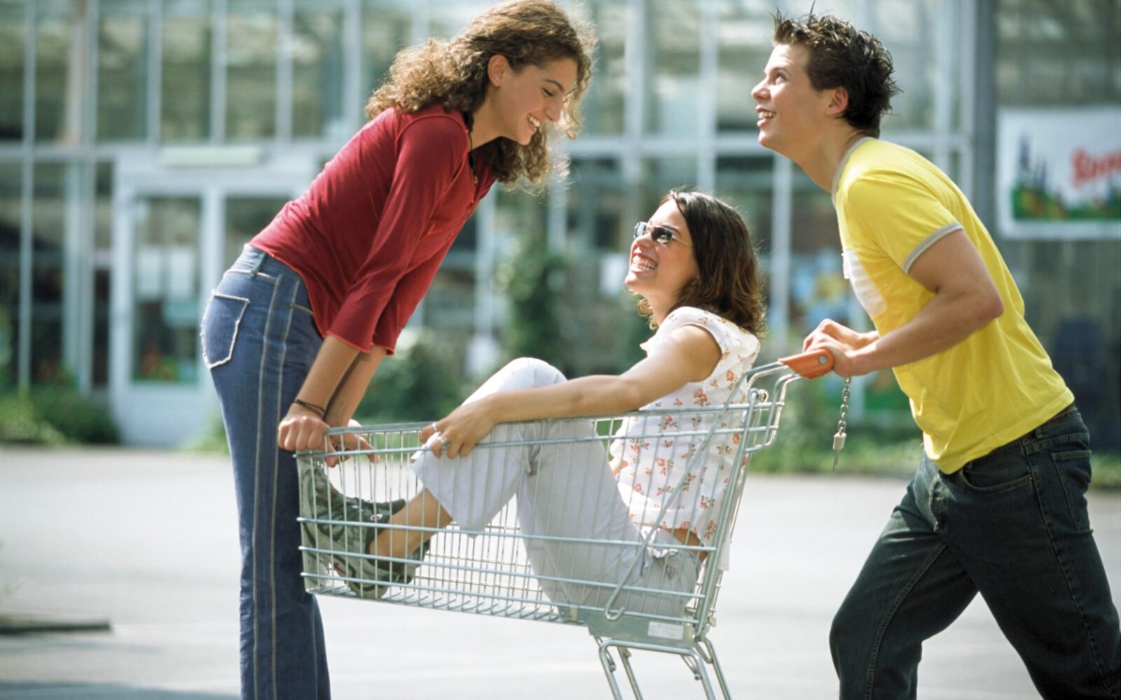 Trije razposajeni najstniki se zabavajo z nakupovalnim vozičkom: ena mladostnica sedi v vozičku, ena stoji na prednji strani, mladostnik pa vozi voziček