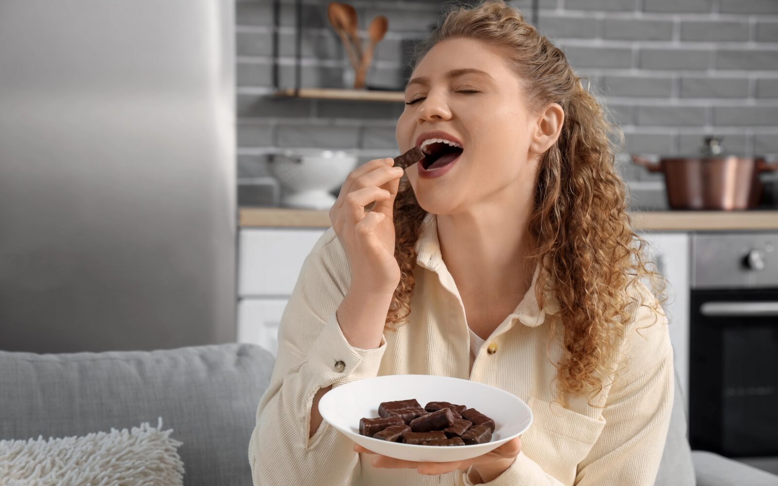 Mlada ženska sedi na kavču in je čokolado - nenaden dvig in padec krvnega sladkorja po neuravnoteževenm obroku