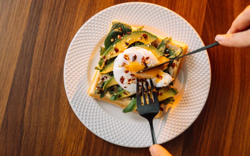 Na krožniku je toast kruh, z rezinami avokada in jajcem na oko. Nekdo je ravno zarezal z vilico in nožem