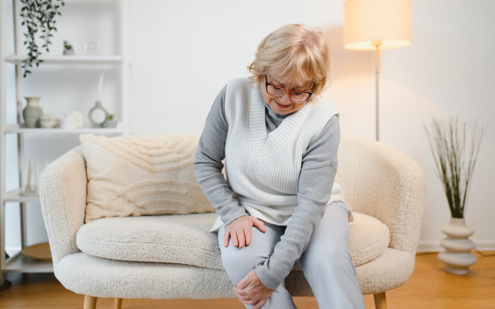 Starejša žensak sedi na kavču in se drži za koleno zaradi bolečin