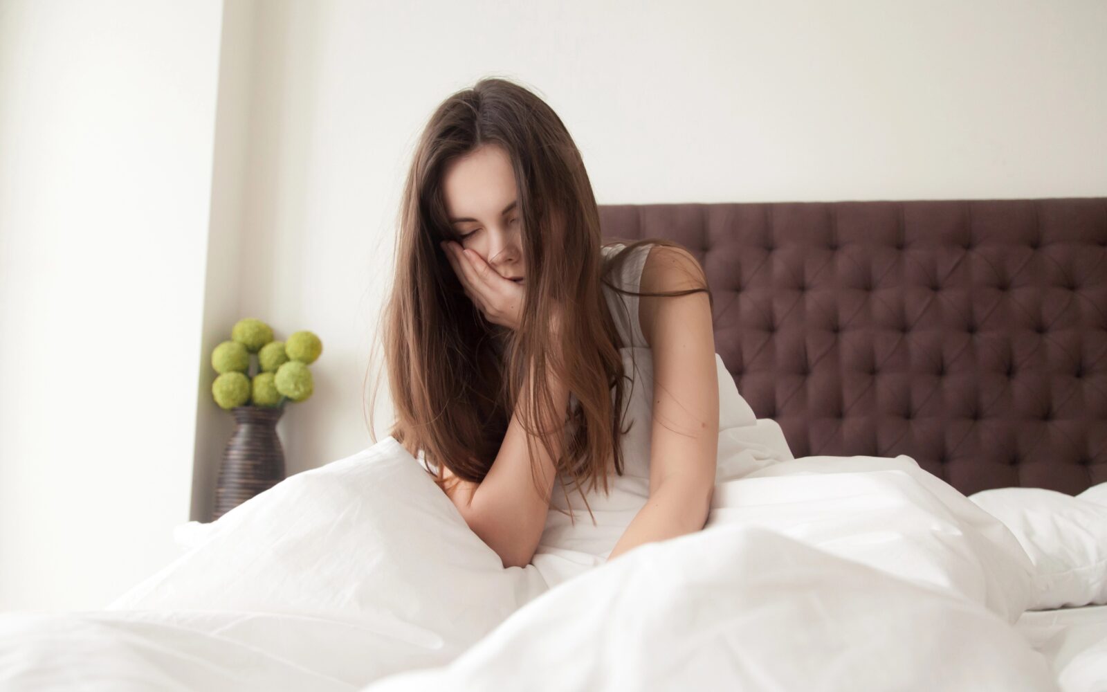 Izmučena in zaspana ženska s skuštranimi lasmi zjutraj miže sedi v postelji