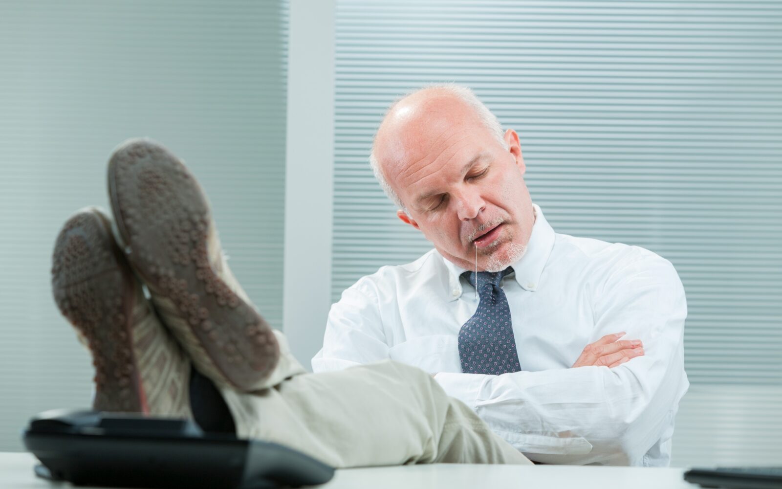 Poslovni moški je zaspal za mizo, noge ima na mizi, iz ust mu teče slina