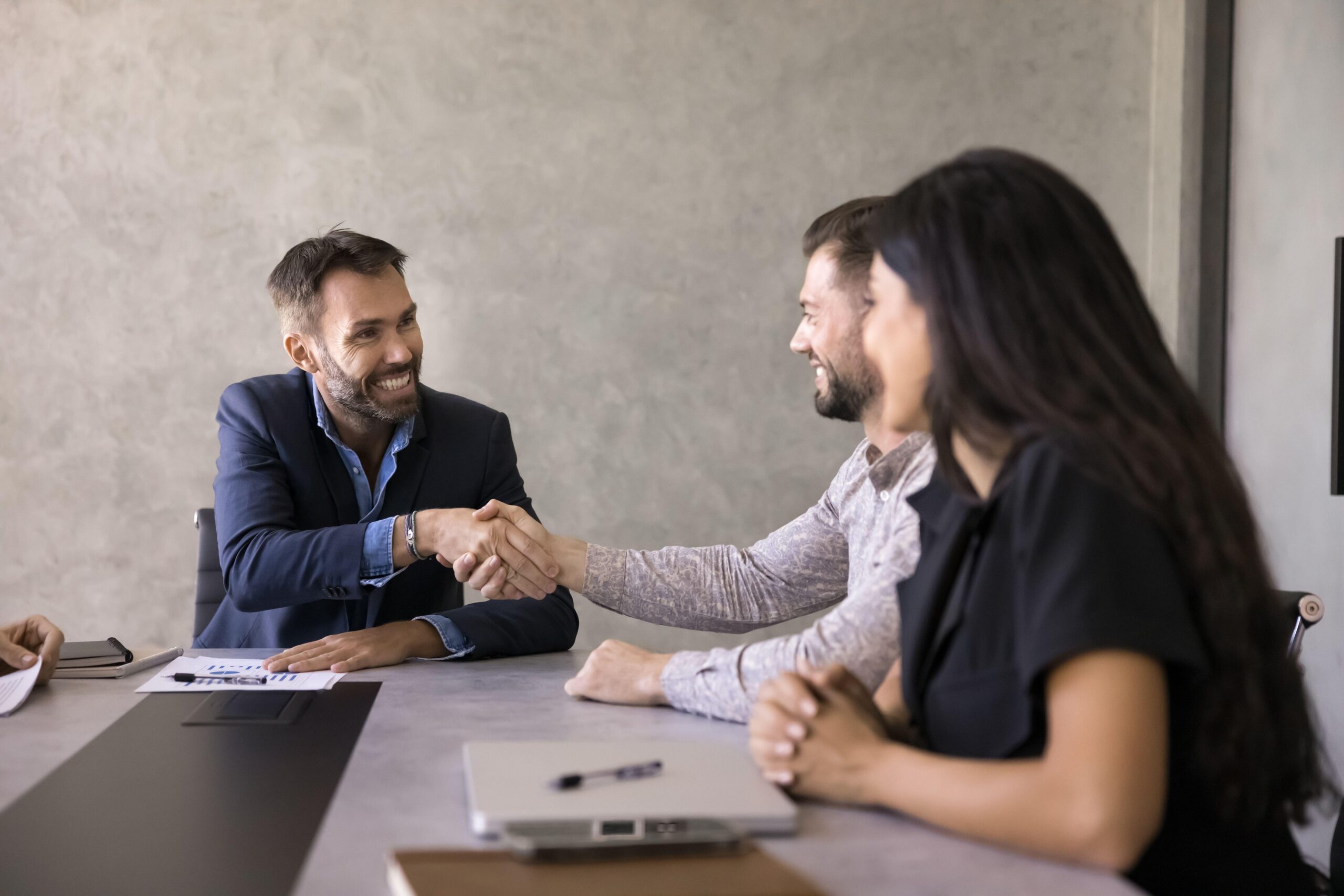 Mladi poslovni ljudje sedijo za mizo na sestanku in se smejijo ter se zadovoljno rokujejo