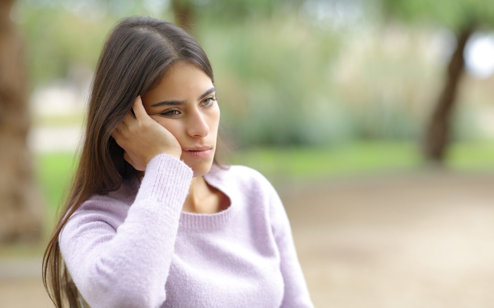 Mlada ženska sama sedi na klopci v parku in izgleda utrujeno - spomladanska utrujenost