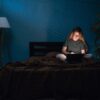 Ženska v temi ponoči sedi na postelji in dela za računalnikom - nočni tip osebe