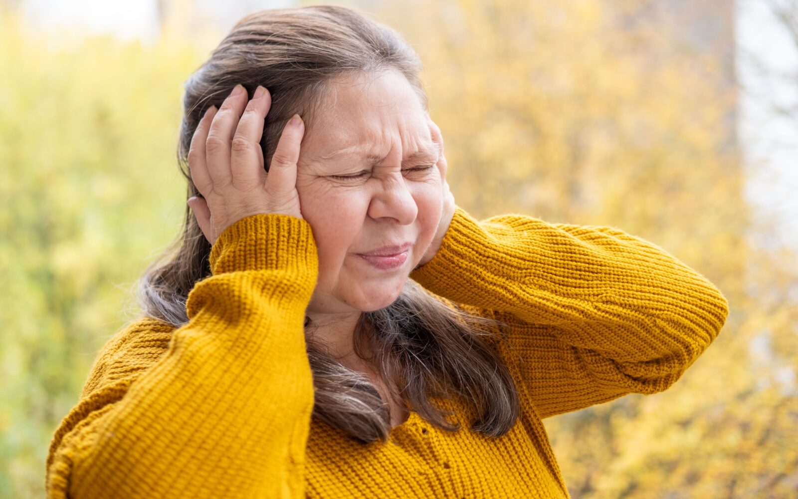 Ženska v rumenem puloverju stoji zunaj in se od bolečine drži za glavo - vzrok je morda v visokem krvnem tlaku