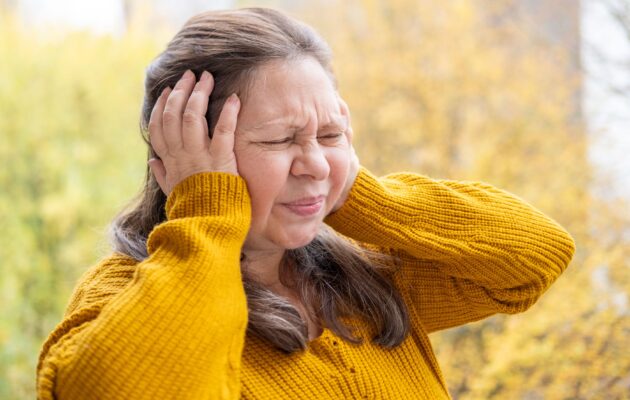 Ženska v rumenem puloverju stoji zunaj in se od bolečine drži za glavo - vzrok je morda v visokem krvnem tlaku