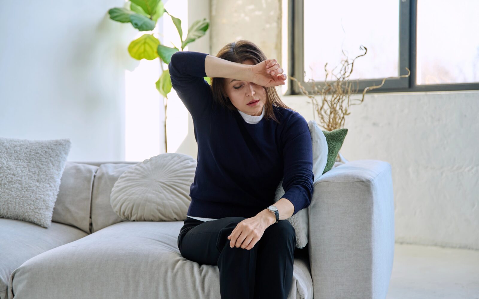 Zrela ženska sedi na kavču in se drži za glavo; je utrujena, nihanje hormonov vpliva na simptome perimenopavze