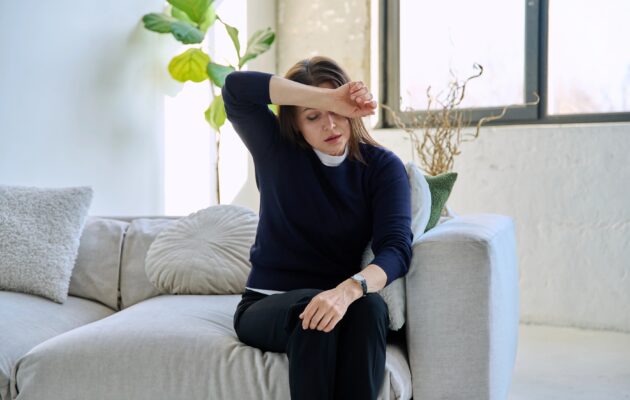Zrela ženska sedi na kavču in se drži za glavo; je utrujena, nihanje hormonov vpliva na simptome perimenopavze