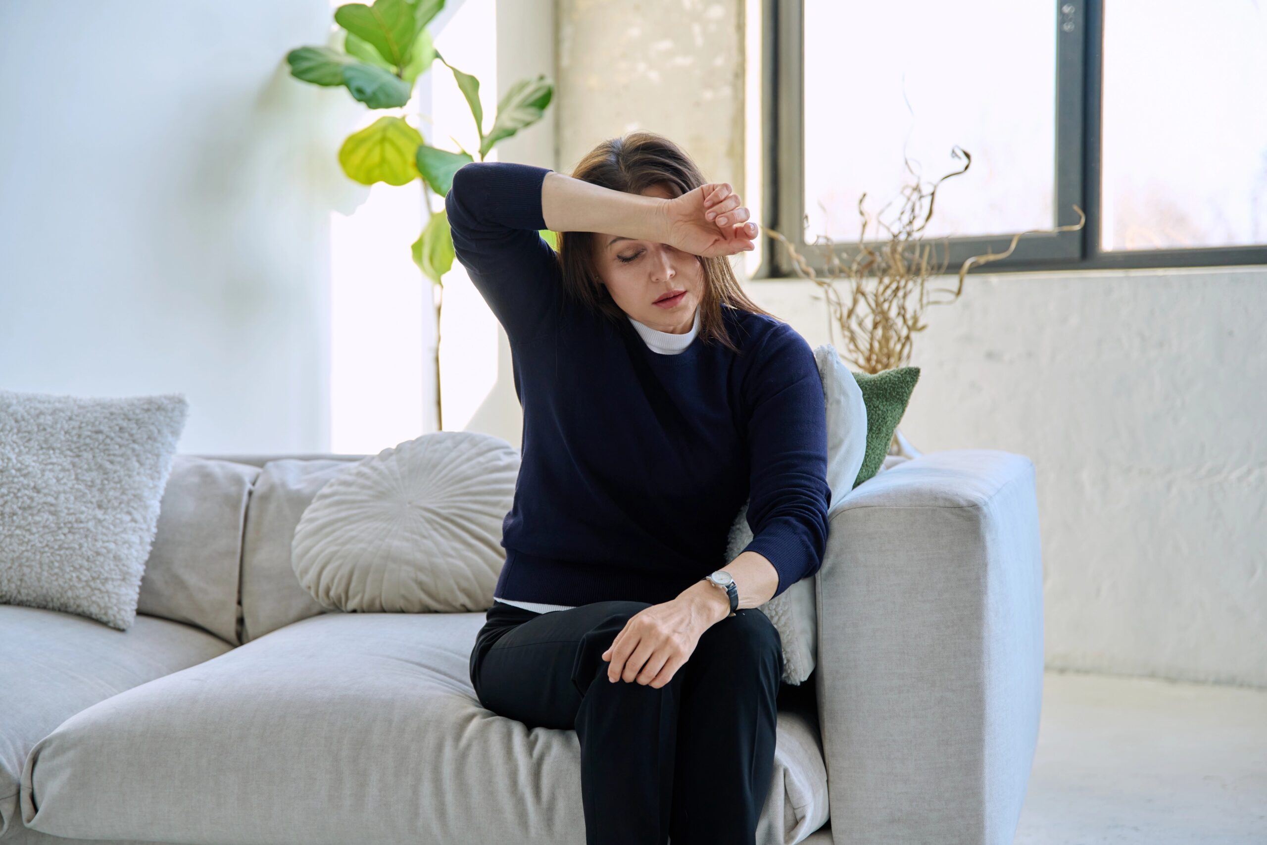 Zrela ženska sedi na kavču in se drži za glavo; je utrujena, nihanje hormonov vpliva na simptome perimenopavze