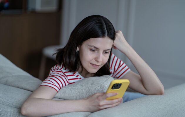 Mlada ženska sedi na kavču in se nasmiha telefonu, ker gleda sproščujoče in pomirjujoče vsebine na socialnih omrežjih