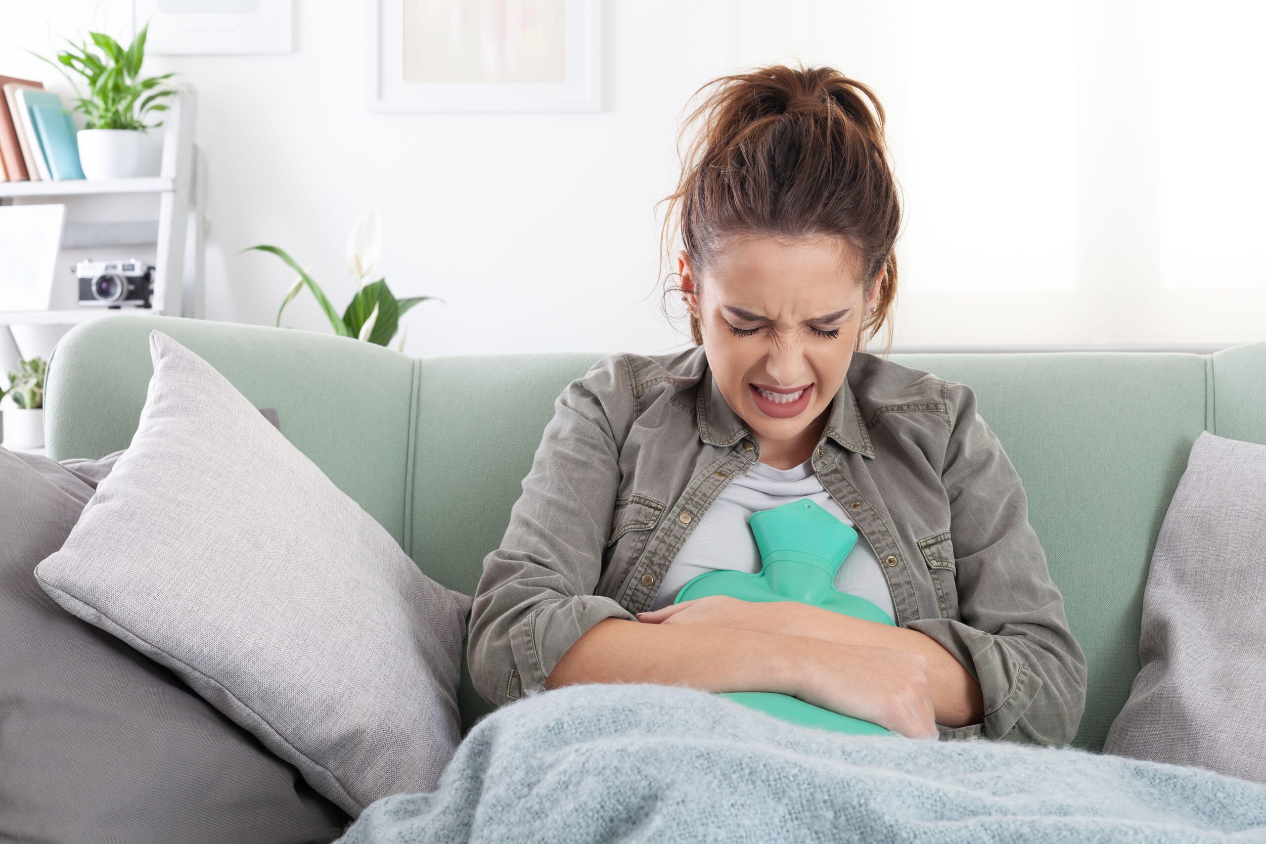 Mlada ženska sedi na kavču in ima na trebuhu termofor za lajšanje bolečin, ki so lahko posledica endometrioze