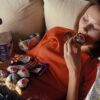 Mlada ženska leži na kavču in je krofe, čokolado, čips in pije sladke pijače - kompulzivno prenajedanje