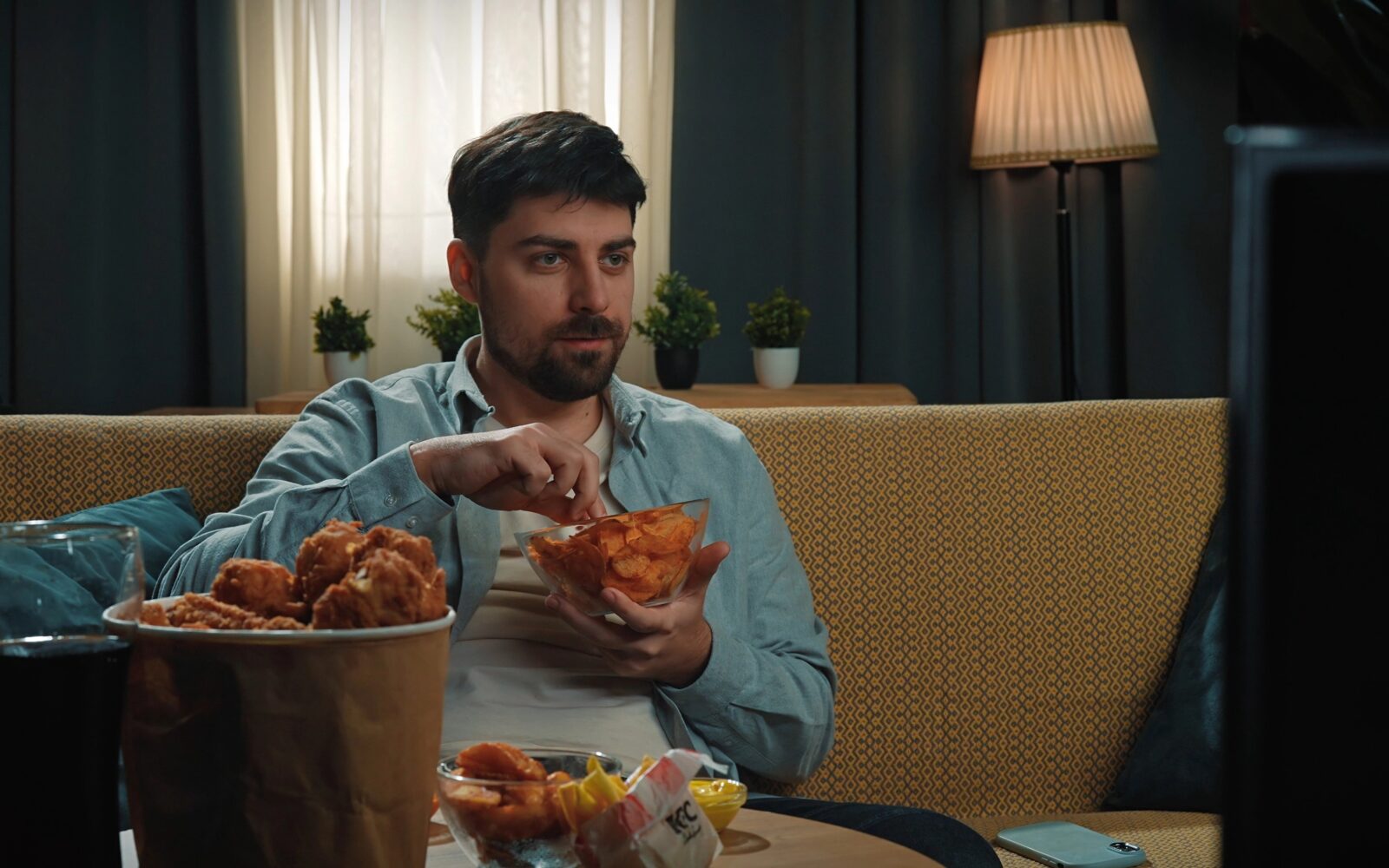 Mlad moški z brado sedi na kavču, gleda v ekran in je hitro, visoko-predelano hrano - čips, omaka, ocvrt piščanec, gazirana pijača