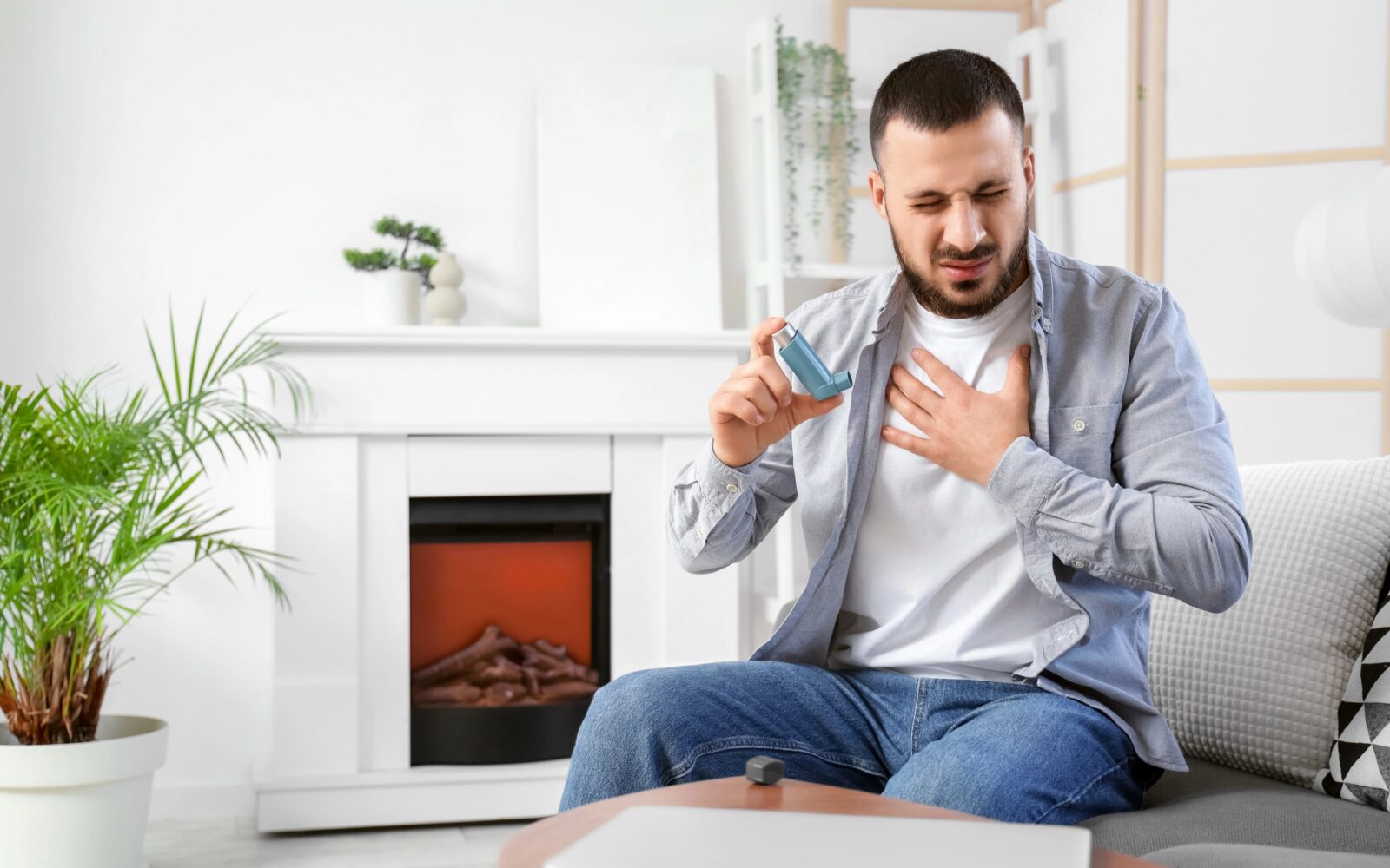 Mlad moški z inhalatorjem v roki sedi na kavču v dnevni sobi in se drži za prsi - ima astmatični napad