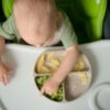 dojenček sedi v stolčku in mu uvajajo gosto hrano, pred seboj ima zelenjavo in jajca