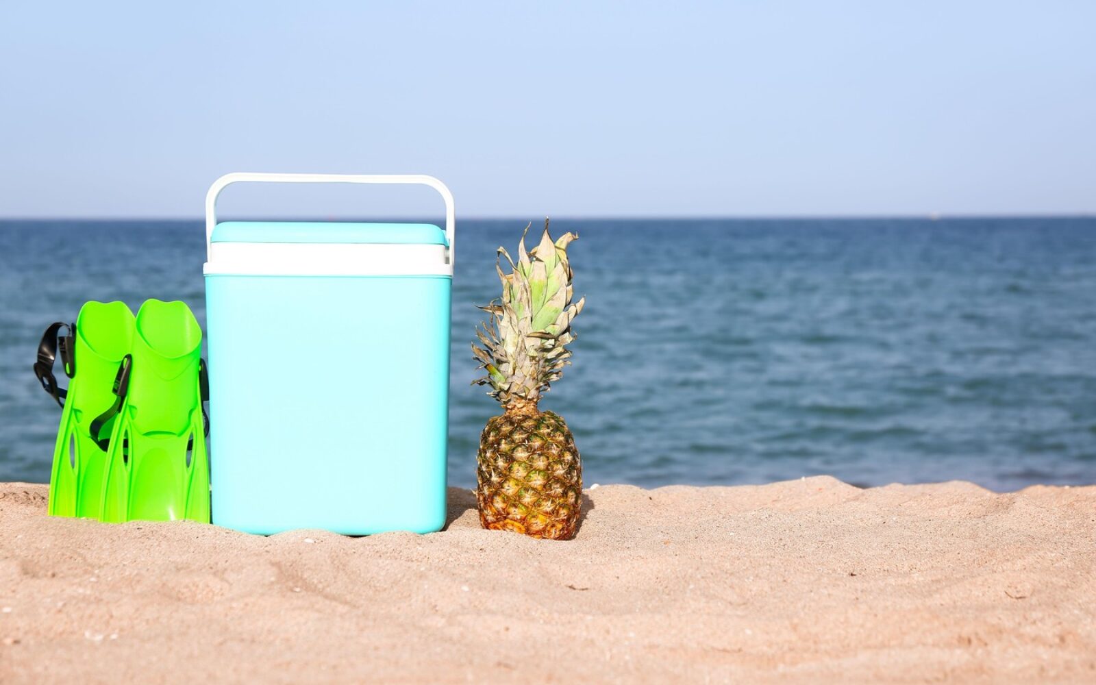 Modra prenosna hladilna torba, zelene plavuti in cel ananas na peščeni plaži ob morju