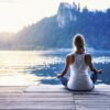 Ženska sedi na pomolu in meditira, v ozadju sta Blejsko jezero, Blejski grad in Zaka