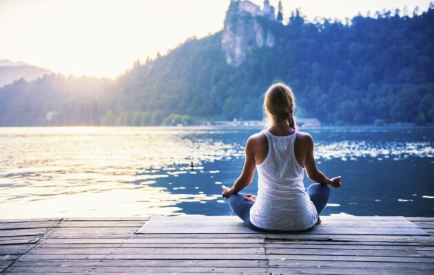 Ženska sedi na pomolu in meditira, v ozadju sta Blejsko jezero, Blejski grad in Zaka
