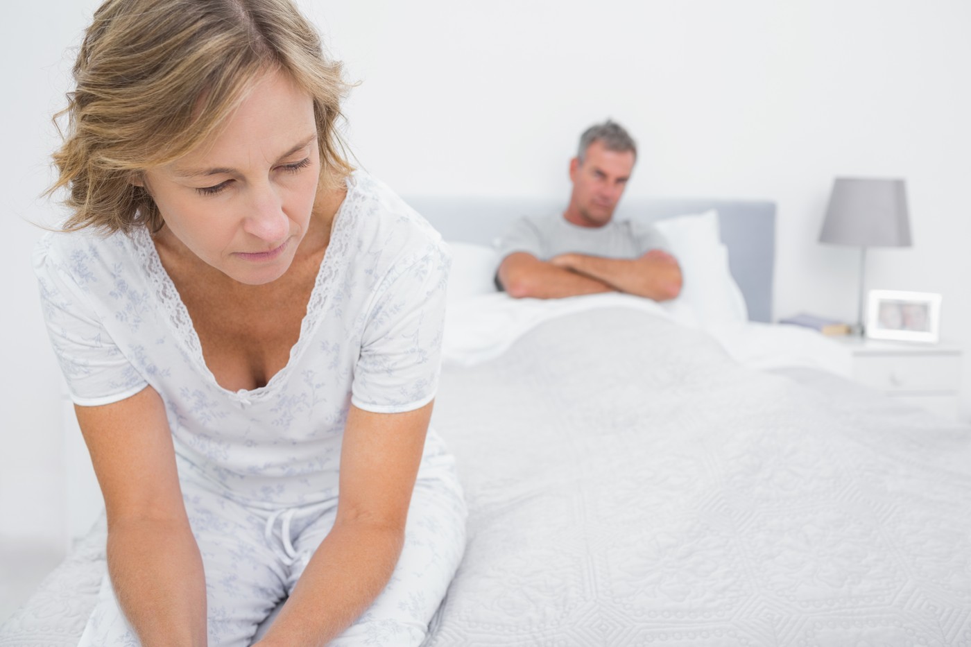 Ženska v menopavzi sedi na postelji, medtem ko partner v ozadju kaže znake odtujenosti. Menopavzni urogenitalni sindrom lahko povzroča boleče spolne odnose in vpliva na partnerski odnos.