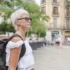 Ženska v beli srajci in sončnih očalih hodi po ulici z nahrbtnikom na hrbtu