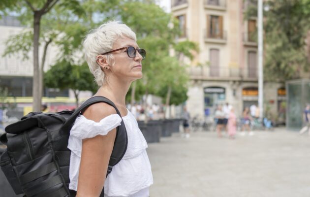 Ženska v beli srajci in sončnih očalih hodi po ulici z nahrbtnikom na hrbtu