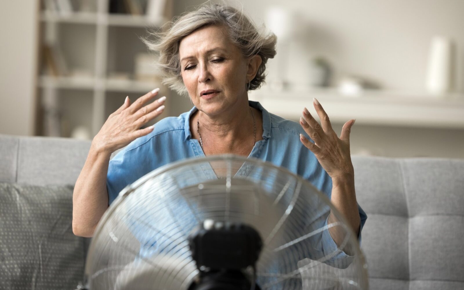 Starejša gospa sedi na kavču in se z rokami hladi pred delujočim ventilatorjem med vročinskim valom doma.