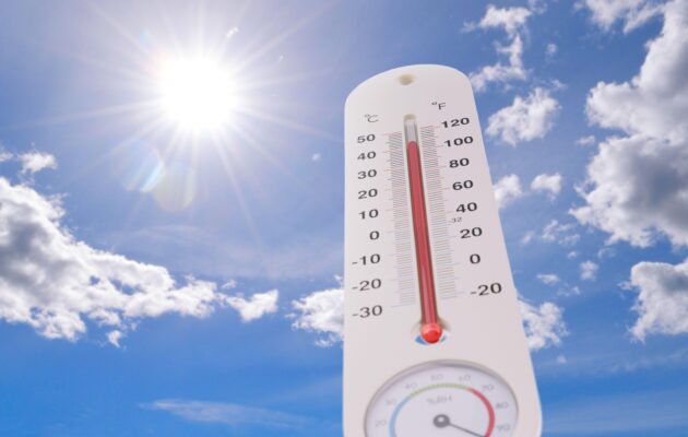 Zunanji termometer prikazuje ekstremno visoke temperature, v ozadju žgoče sonce in modro nebo, ki simbolizirata poletne vročinske valove