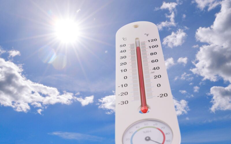 Zunanji termometer prikazuje ekstremno visoke temperature, v ozadju žgoče sonce in modro nebo, ki simbolizirata poletne vročinske valove