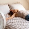 Pregreta mlada ženska leži na postelji in počiva pred ventilatorjem - visoke temperature vplivajo tudi na kakovost našega spanca