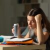 Mlada študentka utrujeno sedi za mizo in se zvečer uči za izpit, v roki ima pločevinko energijske pijače