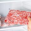 Roka izbira zamrznjeno surovo meso iz zamrzovalnika, kjer je meso shranjeno pri nizki temperaturi za ohranjanje kakovosti.